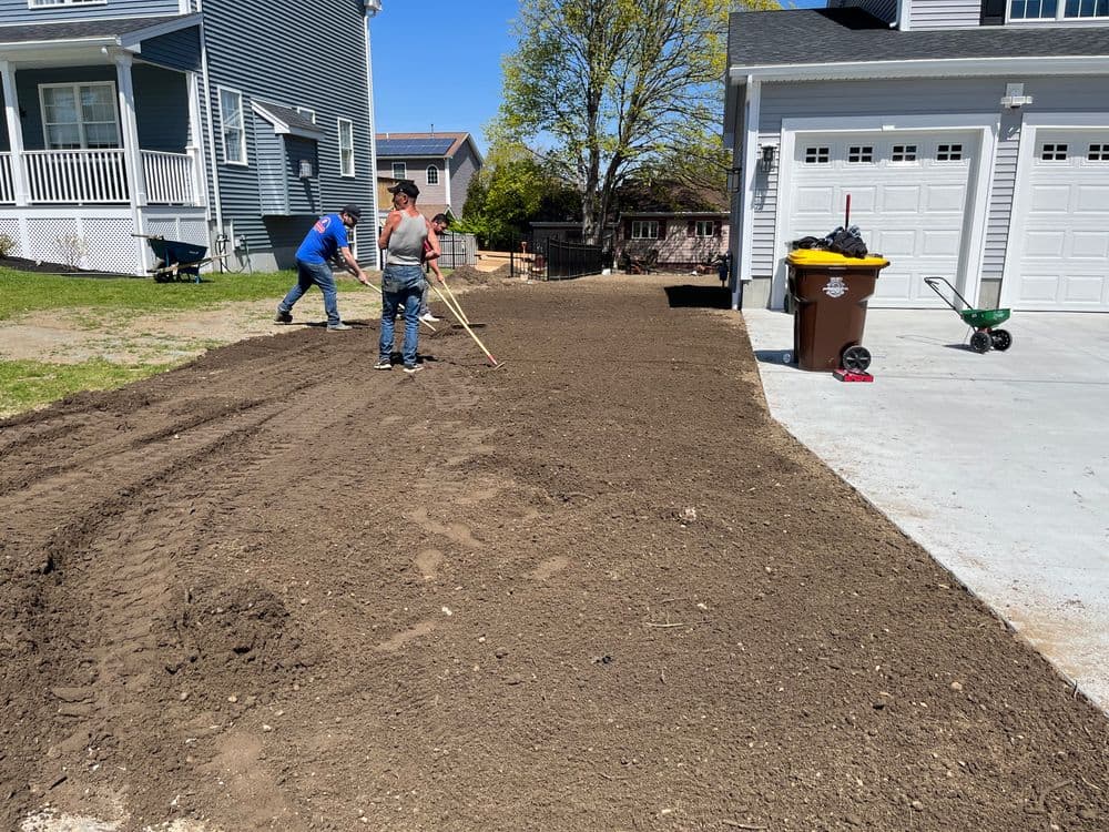 Workers raking and leveling fresh soil in residential driveway during landscaping project