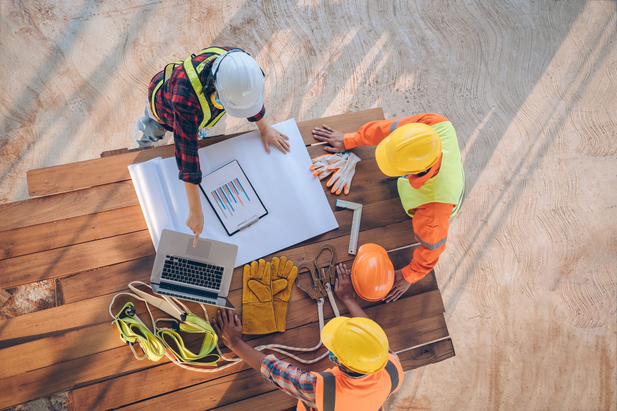 Three construction workers reviewing blueprints and laptop at wooden table with safety equipment